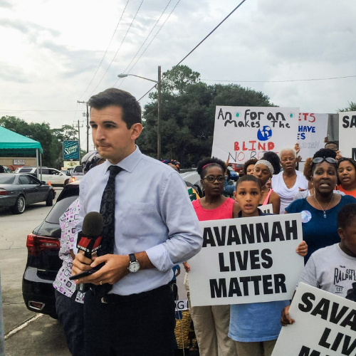 Steven Gallo doing a news broadcast in front of protesters